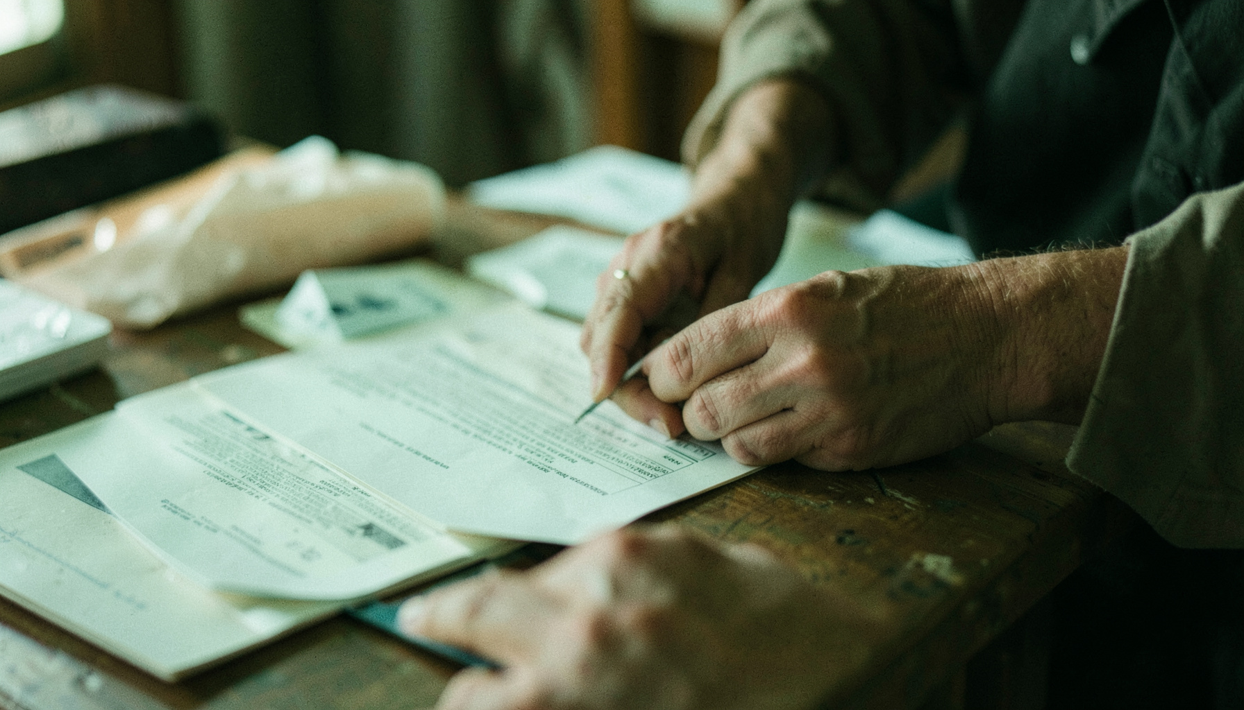 A close-up of hands arranging printed materials on a table, with soft natural light and muted greens and browns.
